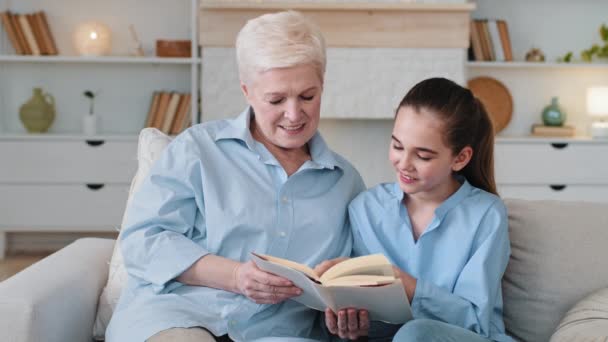 Nieta inteligente leyendo con la abuela en casa. Abuela mayor niñera o tutor privado de edad avanzada ayuda a la colegiala con la tarea explica material se sienta juntos en concepto de pasatiempo sofá — Vídeo de stock