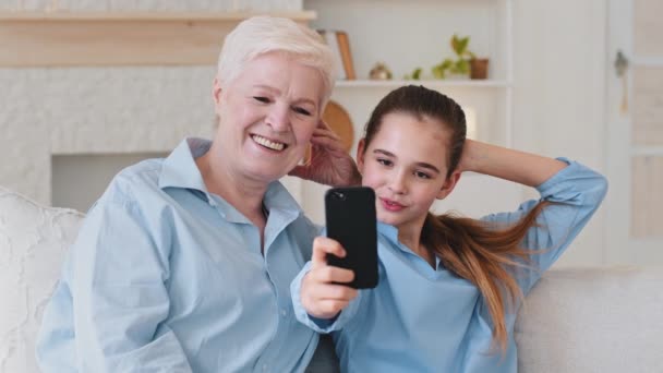幸せな二世代の女性家族シニア祖母と孫娘は携帯電話を見ていますモバイルアプリでセルフィーを楽しみますスマートフォンを持っています面白いソーシャルメディアビデオを見ます自宅で座っているソファ — ストック動画