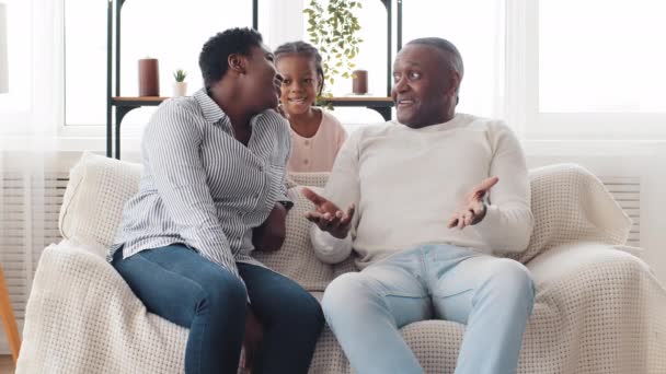 Afro americano familia hablando sentado en sofá en sala de estar, africano mujer madre y maduro negro marido abuelo charlando niña niño hija saltar tocando padres por hombros — Vídeo de stock