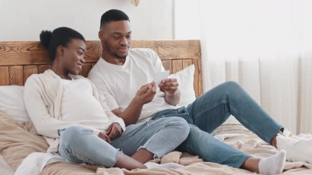 Gelukkig afrikaanse amerikaanse paar getrouwd man en vrouw zwanger afro zwart etnische familie toekomstige ouders zitten op bed thuis genieten van zwangerschap kijken echografie foto 's van ongeboren baby — Stockvideo