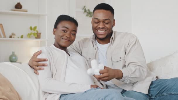 Família afro-americana de futuros pais sentados no sofá em casa marido homem africano pai andando com pequenos sapatos de bebê na barriga grávida de casal esposa amada abraçando amor feliz gravidez — Vídeo de Stock