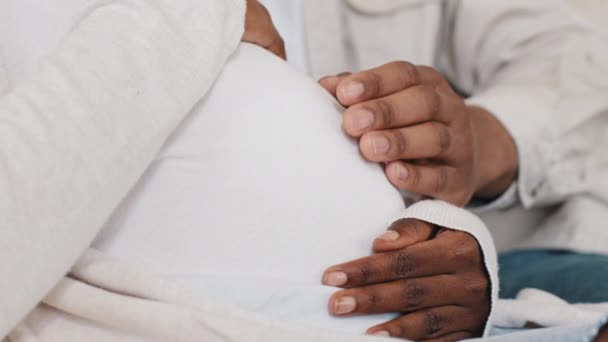 Close-up mãos masculinas e femininas acariciando barriga grávida de mulher em roupas brancas, casal afro-americano irreconhecível futuros pais família desfrutando de gravidez tocando barriga esperando pelo bebê por nascer — Vídeo de Stock