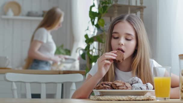 Beyaz liseli kız çocuk ev yapımı çikolatalı bisküvi yiyor portakal suyu ile kahvaltı yapıyor mutfakta oturuyor ve bulanık anne bulaşık yıkıyor. — Stok video
