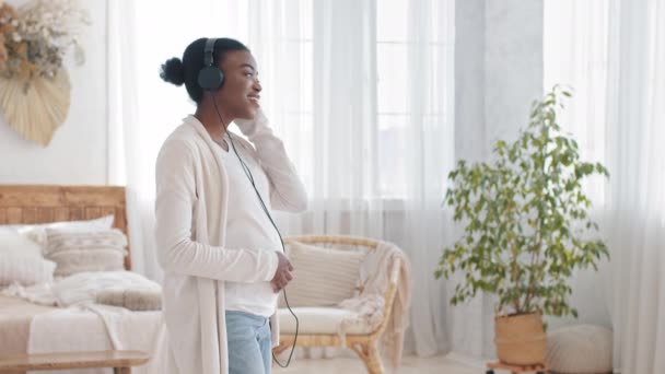 Happy african american expectant mother afro black ethnic woman dancing at home listening to music with headphone enjoy song melody took off put earphone on tummy belly to unborn baby — Stok Video