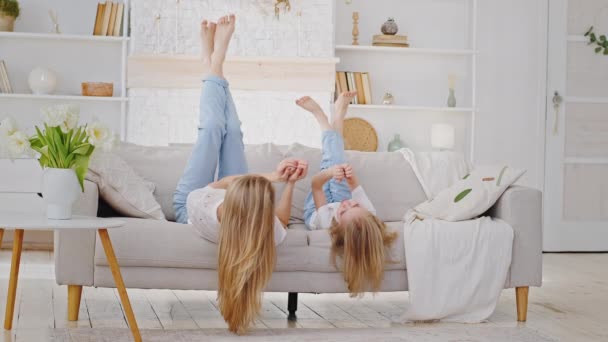 Kaukasische Familie einsame Mutter mit langen Haaren und kleine Mädchen Tochter Kind tragen lässige Kleidung Jeans liegen kopfüber auf dem Sofa spielen berührende Hände reden verbringen Zeit miteinander entspannen drinnen — Stockvideo