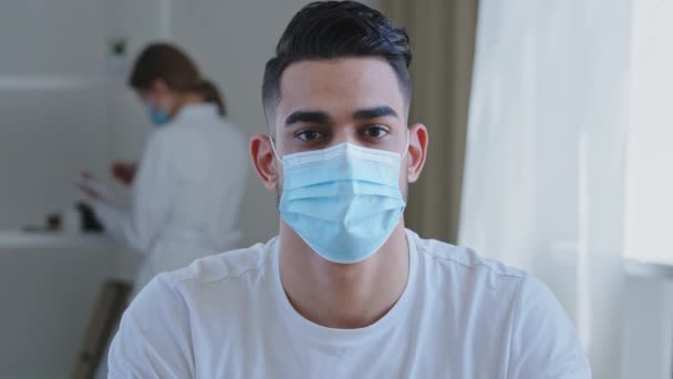 Retrato de hombre árabe étnico enfermo paciente masculino lleva máscara protectora médica en la cara mirando la cámara sentada en la mesa en la clínica en la consulta del hospital en el fondo del médico femenino borroso — Vídeo de stock