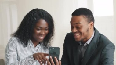 African American millennial students or mixed race businesspeople holding smartphone looking at phone screen, receive good message, great news, excellent result, smiles at camera showing hand OK sign