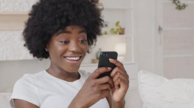 Smiling African American millennial woman holding smartphone having fun playing mobile games enjoy technology online shopping. Happy mixed race lady surfing social media mobile apps content on phone