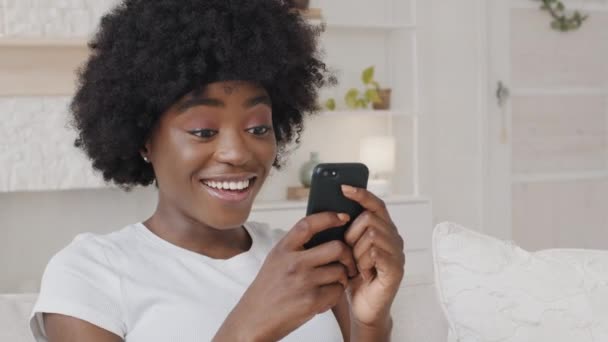 Smiling African American millennial woman holding smartphone having fun playing mobile games enjoy technology online shopping. Happy mixed race lady surfing social media mobile apps content on phone — ストック動画