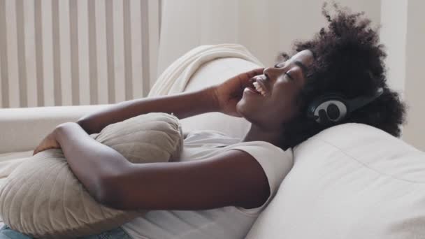Calm smiling African young woman in headphones chilling sitting on sofa with eyes closed listening to favorite slow pacifying music using online player enjoy peaceful mood wearing earphones at home — Vídeo de Stock