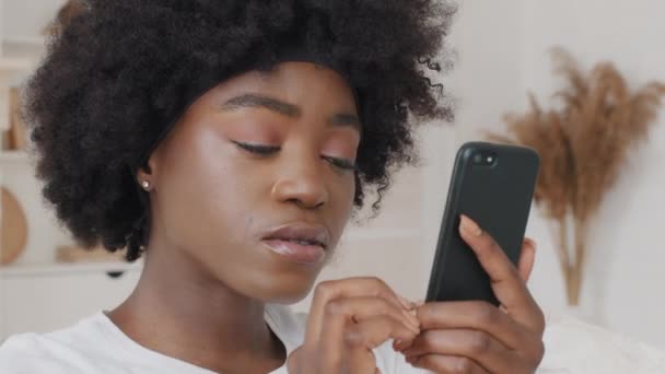 African American woman watching funny photo or video on smartphone at home. Happy lady using apps, reading good news, communication in social network, surfing internet looking at cell sitting on couch — Video Stock