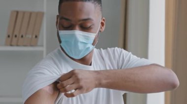 Mutlu genç Afro-Amerikan erkek etnik zenci hasta beyaz tişört giyiyor ve tıbbi koruyucu maske takıyor. 19 numaralı aşıdan sonra kolunda iğne izi var.
