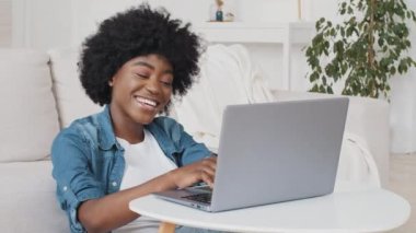 Mutlu Afrikalı Amerikalı genç bir kadın uzaktan kumandayla uzaktan kumandayla gülümsüyor. Kadın vaktini sosyal ağlarda sohbet ederek geçirir. Laptop kullanır, iş veya öğrenim için yararlı bilgiler arar.