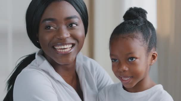 Портрет щасливої афро-американки, чорної радісної матері, яка дбайливо обіймає африканську маму, обіймає маленьку школярку, гарненьку дівчинку, яка сміється з щирого зубного посмішку, сидячи разом у приміщенні. — стокове відео