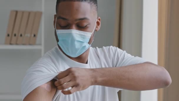 Felice giovane afroamericano uomo etnico nero ragazzo paziente indossa bianco t-shirt e medico maschera protettiva mostra segno di iniezione sulla toppa spalla sul braccio dopo la vaccinazione covid19 fa come pollice in su — Video Stock