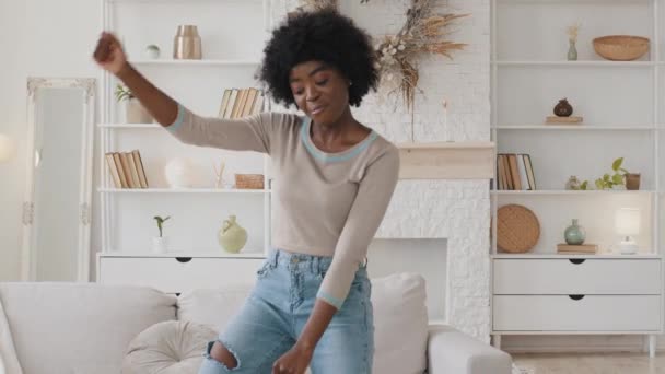Χαρούμενη ανέμελη νεαρή Αφρο-Αμερικανίδα που χορεύει ακούγοντας καλή μουσική στο σπίτι μόνη της. Αξιολάτρευτο ενεργειακό millennial μικτή φυλή κορίτσι που διασκεδάζουν με στο σαλόνι απολαμβάνοντας ξεκούραση την Παρασκευή — Αρχείο Βίντεο