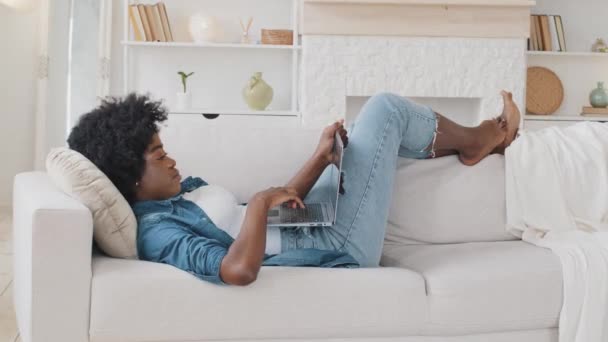 Sonriente joven africana serena acostada en el sofá mensajes de texto en el ordenador portátil. Mujer bastante relajada descansando con gadget en la acogedora sala de estar viendo fotos o videos divertidos, pasando tiempo libre en casa — Vídeos de Stock