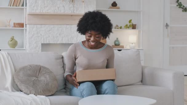 Lachende Happy African jonge vrouw klant zitten op de bank shakes pakket doos tevreden met online winkel bestelling levering thuis, glimlachende consument ontvangt goede aankoop per post verzendconcept — Stockvideo