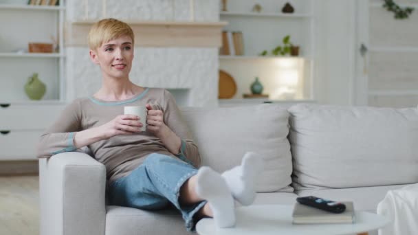 Dreamy beztroska szczęśliwa kobieta siedzi na kanapie pijąc poranną kawę lub ciepłą herbatę w domu, uśmiechnięta atrakcyjna pani trzyma filiżankę relaksu czuje się bezstresowo korzystając przytulny styl życia na przyjemny weekend — Wideo stockowe