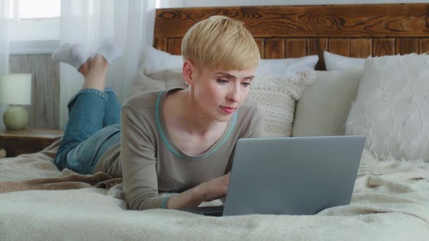 安详镇定的女人一个人躺在床上休息。她在社交媒体网络上使用手提电脑观看视频，在网上播放电视连续剧。成年女士在周末懒洋洋地运用现代技术概念 — 图库视频影像