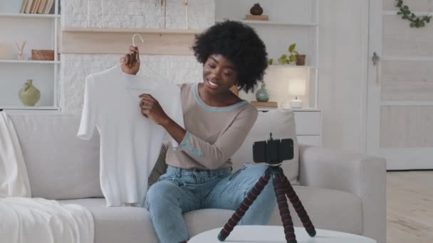 Młoda afrykańska blogerka siedząca na kanapie nagrywająca wideo przed kamerą, dzieląca się doświadczeniami zakupowymi z wyznawcami. Millennial robi transmisję na żywo dla kanału social media reklamy biały t-shirt — Wideo stockowe