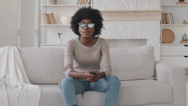 Joyeux jeune femme afro-américaine portant des lunettes 3D assis sur un canapé confortable, changer de canaux à l'aide d'une télécommande, choisir des programmes ou des spectacles, souriant profiter week-end à la maison, passe-temps de loisirs — Video