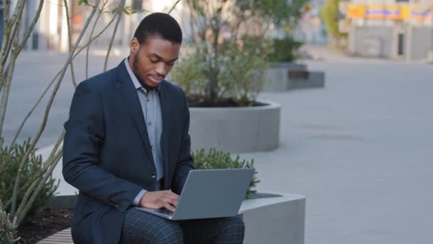 Estudiante afroamericano freelancer enfocado en traje sentado al aire libre sosteniendo portátil en el estudio de rodillas, escribiendo en el ordenador portátil. Serious joven negro hombre trabajando a distancia en línea prepararse para el examen de prueba — Vídeos de Stock