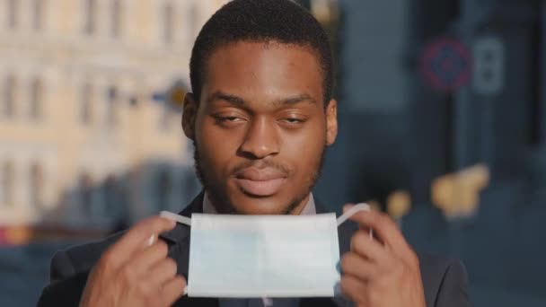 Retrato de headshot Empresário afro-americano coloca máscara médica protetora em pé ao ar livre contra alto fundo do edifício olhar para a câmera. Parar o surto de coronavírus, conceito de contaminação do ar — Vídeo de Stock