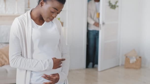 Cortado tiro grávida africano jovem mulher étnica grávida mãe acariciando barriga afro preto carinhoso marido futuro pai entra no quarto traz vidro de laranja fruta saudável suco fresco abraços esposa — Vídeo de Stock