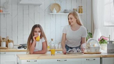 Caucasian mother mommy puts plate of cookies on table hugging cuddling talks with beloved daughter girl drinking orange juice smiling together standing in home kitchen enjoy hug, family love concept
