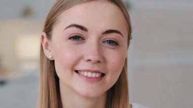 Cheerful pretty millennial single woman looking smiling at camera in living room indoors. Happy young adult beautiful lady with attractive face closeup portrait. White healthy teeth, dentistry concept