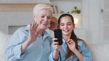 Old grandmother and little granddaughter having fun using smartphone look at screen laugh talk make online video call take selfie bonding sit on sofa at home. Two generation family with gadget concept