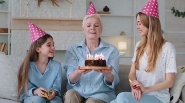 Happy excited beautiful three women age generations family old grandmother young mom and little child granddaughter celebrating birthday in party hats congratulations together. Grandma blowing candles
