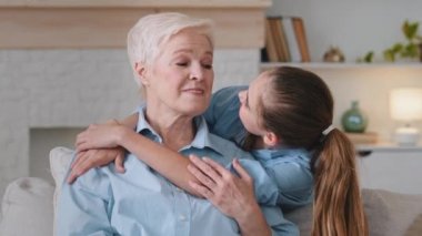 Sad, serious elderly lady mature senior sits relaxing on cozy sofa at home alone. Little cute loving granddaughter hugs grandmother, laugh, rejoice, enjoy weekend together. Caring for elderly concept