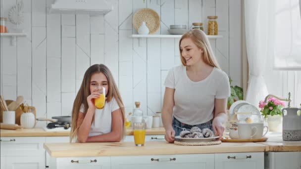 Caucasian mother mommy puts plate of cookies on table hugging cuddling talks with beloved daughter girl drinking orange juice smiling together standing in home kitchen enjoy hug, family love concept — Stok video