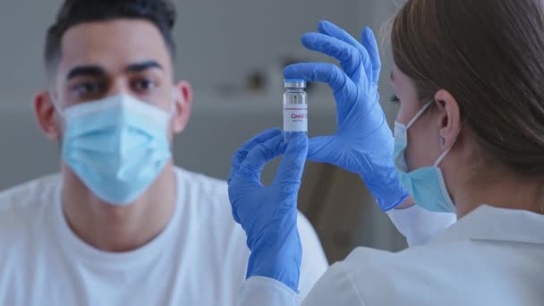 Widok z tyłu kaukaska kobieta lekarz pielęgniarka farmaceuta naukowiec nosi maskę medyczną i ochronne rękawice lateksowe posiada butelkę szczepionki przeciwzakrzepowej w rękach koronawirus pokazuje do zamaskowanego mężczyzny — Wideo stockowe