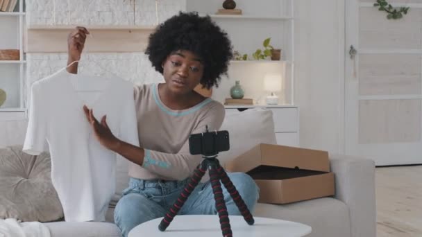 Glückliche junge afrikanische schöne Bloggerin, die vor dem Handy auf einem Stabilisator sitzt und Modebekleidungsvideos für ihren persönlichen Kanal aufnimmt. Lächelnde Influencerin teilt Einkaufserlebnis — Stockvideo