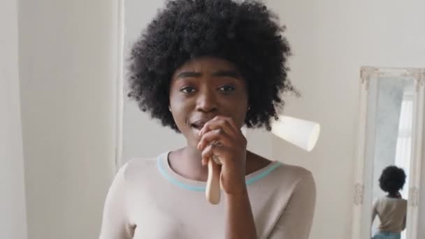 Happy African American millennial young black woman listening music sing along into hairbrush enjoying popular rhythmic song playlist relaxing in living room. Stress-free home weekend rest concept — Stok video