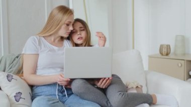 Serious mother Caucasian business woman customer sitting on couch with teen daughter looking at laptop choosing goods online watching movie browsing child girl yawning falls asleep on shoulder of mom