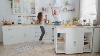 Funny adult mother happy girl teenager daughter child holding hands jumping dancing whirling in kitchen at home playing with long hair moving to music enjoy weekend dance celebrate having fun together