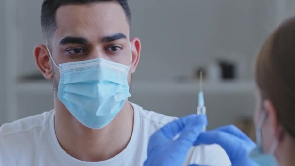 Detailní záběr arabský Španěl v lékařské masce sedí na klinice a připravuje se na vakcinaci a dívá se na nerozpoznatelnou sestru lékaře s injekční stříkačkou s vakcínou covid-19 lék — Stock video