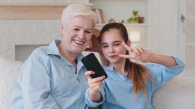 Funny elder mature grandma and little granddaughter bloggers having fun video calling on smartphone shoot social media video, happy grandmother laugh look at cellphone using app sit on sofa at home