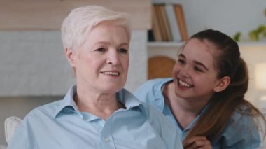 Happy hoary mature senior relaxing on comfortable sofa. Adorable granddaughter plays, covering eyes with palms grandmother. Smiling small kid girl and pleasant happy granny having fun together home