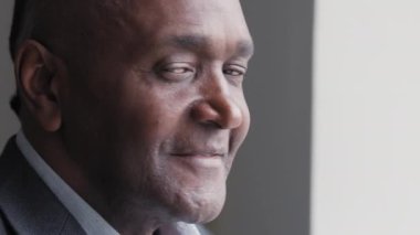 Male portrait of african american mature afro man with pensive dreamy expression on face wrinkled boss leader looking out window in office dreaming resting planning looking at camera winking smiling