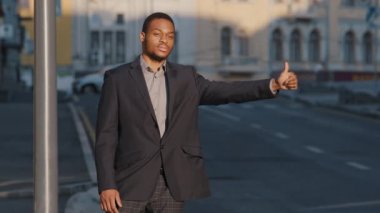 Confident calm mixed race young adult businessman wearing suit raising hand showing thumb up outdoors. African American black man standing on sidewalk by road trying to get cab in morning or evening
