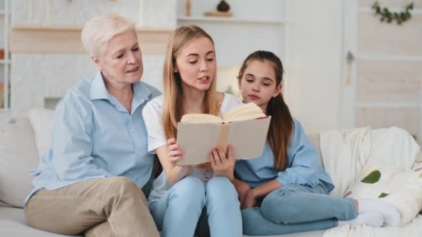 Disparo en la cabeza feliz amante mujer de mediana edad y nieta hija pequeña escuchando adulto sonriente hija y madre leyendo libro de cuentos de hadas de papel. Familia femenina se sienta en sofá pasar fin de semana en casa — Vídeos de Stock