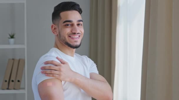 Happy satisfied ethnic guy arab man hispanic patient male posing indoors wears white t-shirt shows plaster on shoulder feels relief joy after vaccination injection with covid remedy medical procedure — Αρχείο Βίντεο