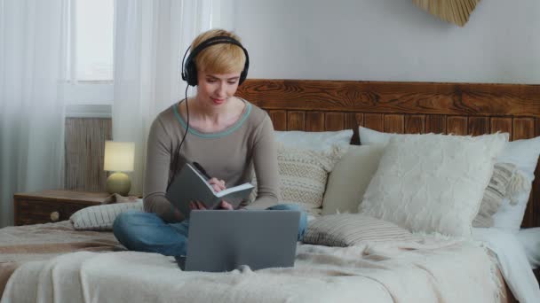 Smilende vakker kvinne i hodetelefoner blir hjemme mens hun ser på laptop-skjermen, bruker video-call-app mens hun ser på forelesninger om utdanning, skriver notater i en bok, sitter på en komfortabel seng – stockvideo