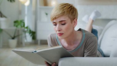Relaxed middle-aged woman lying on comfy couch in living room reading new favorite paper book novel bestseller, enjoying hobby, leisure time spending cozy weekend at home during cold winter season