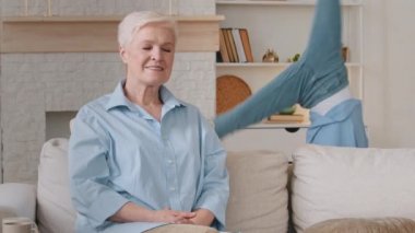 Elderly hoary grandmother sits on couch, smiling little gymnast granddaughter having fun, doing cartwheels, hugs grandmother. Women of two generations rejoice, enjoy spending time together, feel happy